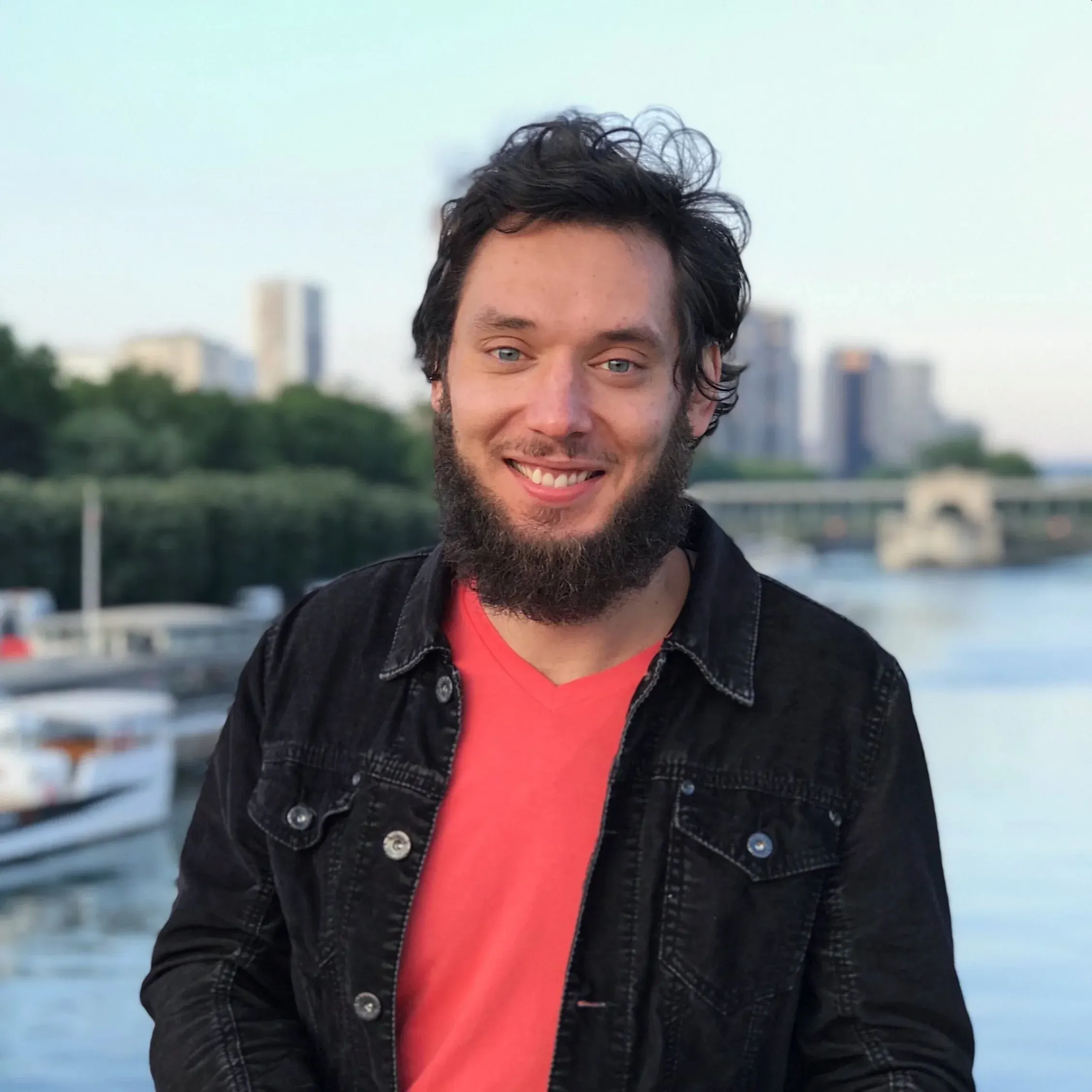 Pavel Novel, smiling, in a black denim jacket and coral T-shirt, by a river with moored boats and a bridge behind him.